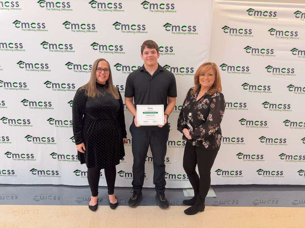 three people standing in front of a backdrop that says CMCSS. Woman wearing all black, male student with a certificate and female in a floral print shirt.