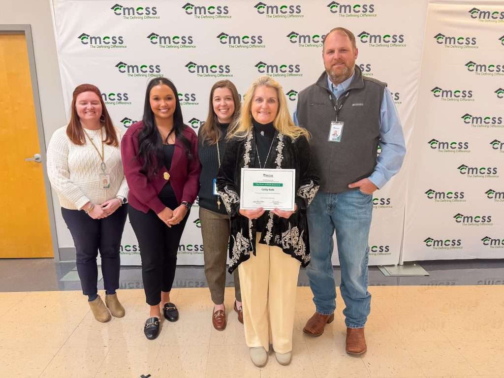 Group of five adults, four female and one male, standing in front of a CMCSS backdrop. Female with blond hair wearing a black sweater and cream colored pants is holding a certificate.
