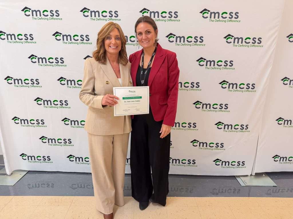 Two women standing in front of a CMCSS backdrop. Woman in a cream colored pant suit is holding a certificate.