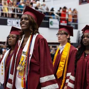group of four graduates walking into room wearing maroon gowns smiling